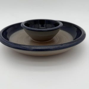 May include: A ceramic chip and dip serving set. The set features a round, shallow dish with a raised rim and a smaller, circular bowl in the center. The rim and the interior of the small bowl are dark blue, while the rest is tan.