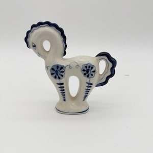May include: A ceramic horse figurine in white with blue accents. The horse has a stylized design with a blue mane, tail, and floral patterns. The figurine is standing on a small base and has a whimsical, decorative appearance.