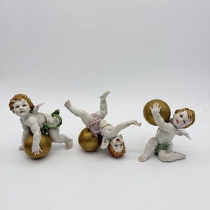 May include: Three porcelain cherub figurines. Each cherub has wings and is posed with a golden sphere. The cherubs have fair skin, red hair, and are wearing light-coloured cloth. One cherub is upside down, another is kneeling, and the third is crawling.
