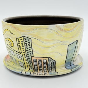 Puede incluir: Un cuenco de cerámica con un diseño de paisaje urbano. El cuenco presenta un fondo amarillo con contornos negros de edificios y un interior marrón oscuro. La base es amarilla con un diseño ondulado.