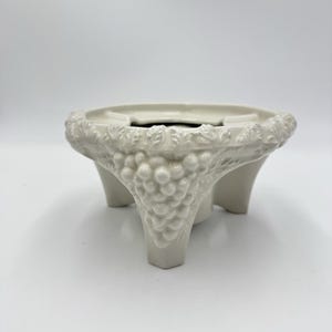 May include: A cream-colored ceramic stand featuring a circular top and three legs. The exterior surface is decorated with a raised, embossed relief pattern of grape clusters and vine leaves that extends from the rim down to each of the three legs.