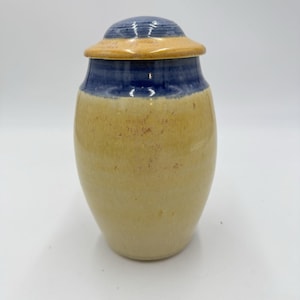 May include: A ceramic jar with a rounded lid, featuring a glossy finish. The body is a light tan color with subtle speckles, while the upper section and the lid are a deep blue. The lid also features a band of tan glaze around its lower edge.