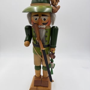 May include: A wooden nutcracker in the form of a hunter, wearing a green hat and jacket, tan shorts, and holding a rifle. The nutcracker has a white beard, a detailed face, and is standing on a wooden base. A decorative pinecone is attached to the hat.