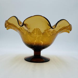May include: An amber-colored glass bowl featuring a distinct crackle texture throughout the body. The bowl is supported by a short pedestal base and has a wide, ruffled, undulating rim that creates a decorative, wavy edge profile.