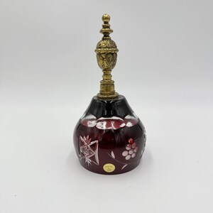 Può includere: Una campana in cristallo tagliato rosso intenso con intricati motivi a stella e a grappolo d'uva. La campana è sormontata da un'impugnatura in ottone testurizzato e decorato e presenta un piccolo adesivo circolare sul lato.