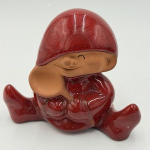 May include: A ceramic figurine of a seated character wearing a glossy, deep red hat and matching outfit. The figure has a matte, unglazed tan face with a simple curved line for a closed eye and a small nose, holding a tan spoon near its face.