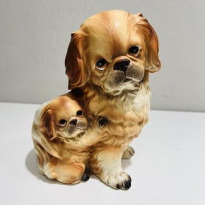 Könnte beinhalten: Eine Keramikfigur eines braunen und weißen Pekinesen mit einem kleineren Pekinesen-Welpen, der sich an ihn kuschelt.