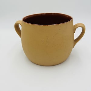 Könnte beinhalten: Eine beige Keramik-Suppenschüssel mit zwei Griffen. Das Innere der Schüssel ist dunkelbraun. Die Schüssel hat eine abgerundete Form und steht auf einer weißen Oberfläche.