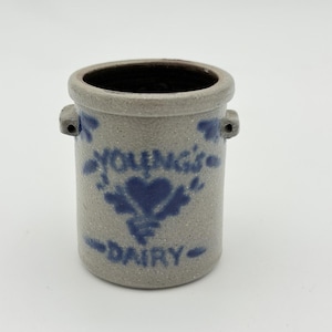 Puede incluir: Un pequeño tarro cilíndrico de gres con base gris y letras y diseño azul oscuro. El texto dice "YOUNG'S DAIRY" con un diseño en forma de corazón. El tarro tiene dos asas pequeñas y un interior oscuro.