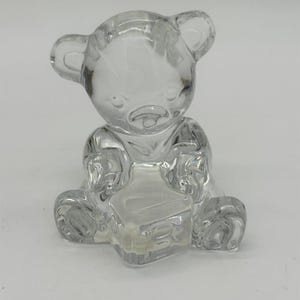 Puede incluir: Una figura de cristal transparente de un oso de peluche sentado que sostiene un bloque cuadrado en su regazo. El oso tiene orejas redondeadas, rasgos faciales definidos y una superficie lisa y reflectante. El bloque tiene la letra B al frente.