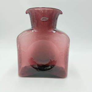 Puede incluir: Un jarrón de cristal color amatista de Blenko Handcrafts. El jarrón tiene una superficie texturizada y una base cuadrada que se curva en un cuerpo redondeado. La parte superior se ensancha con una amplia abertura. Una pequeña etiqueta está adherida al cuello.