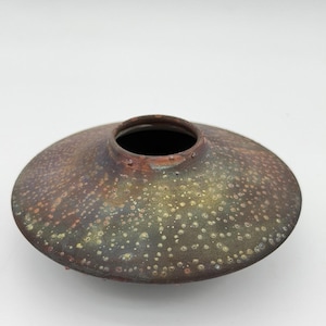 May include: A low-profile ceramic vase with a wide, flattened circular body and a small central opening. The surface features a mottled, textured glaze in shades of dark brown, muted green, and copper, with small, light-colored speckles throughout.