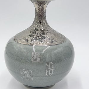May include: A celadon green ceramic vase with a crackle glaze finish. The neck and shoulder feature an intricate silver-toned metallic overlay with floral motifs. The body of the vase is decorated with three distinct white square and oval seal stamps.