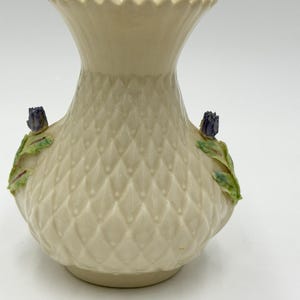 Puede incluir: Jarrón de cerámica color crema con un patrón de diamantes y borde ondulado. El jarrón presenta dos capullos de flores moradas con hojas verdes. Una pieza decorativa para la decoración del hogar.