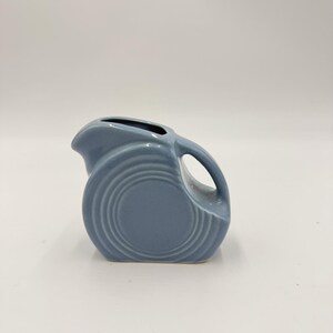 May include: A blue ceramic jug with a rounded body and a curved handle. The jug has a spout for pouring and concentric circle detailing on the front. The jug is a solid blue colour and sits on a white surface.