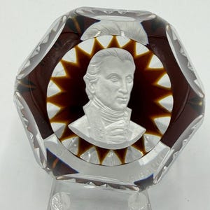 May include: A clear, faceted glass paperweight with a portrait of a man in a white relief. The portrait is surrounded by a brown starburst design. The paperweight has a clear base.