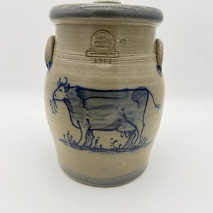 May include: A beige stoneware crock with a lid, featuring a blue hand-painted cow illustration. The vessel has two side handles and a stamped mark reading Handmade Rowe Pottery Works Cambridge Wis 1992. The rim and base have a dark grey glaze.