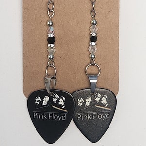 May include: A pair of dangle earrings featuring black guitar picks with a white image of the band Pink Floyd and the band's name. The picks are attached to silver chains with clear and black beads.