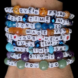 May include: A stack of beaded bracelets with white letter beads spelling out phrases like "OVER FEAR" and "HAVE THE DAY." The bracelets feature various colored beads, including blue, orange, purple, and green, with some gold glitter beads.