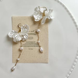 May include: Delicate white floral earrings with gold-tone chains and pearl drops. The earrings are displayed on a small card with text, presented on a white plate with a beaded edge. The earrings have a feminine design, suitable for special occasions.