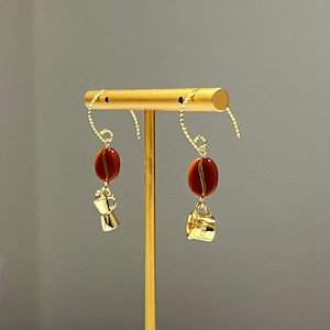 May include: Gold-colored dangle earrings featuring a coffee bean and a miniature coffee maker charm. The earrings have a gold-colored hook and a large, oval, reddish-brown bead. The coffee maker charm is gold-colored.