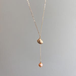 May include: A gold-toned lariat necklace featuring a delicate chain, a small ridged seashell charm, and a single round, pale pink pearl pendant suspended at the bottom of the vertical chain drop, creating a simple and elegant jewelry piece.