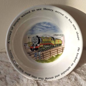 May include: A white ceramic bowl featuring a central illustration of a green steam locomotive numbered 3, with two figures standing by a fence. The rim is inscribed with the text: A crowd of people waited to see Henry arrive in his new shape. He looked so splendid and strong that they gave him three cheers.