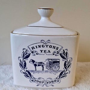May include: White ceramic tea caddy with a lid and gold trim. The front features a blue illustration of a horse-drawn carriage, the text "Rington's Tea", and the years "1907-1982".
