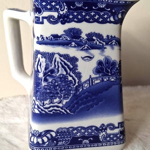 May include: A white ceramic pitcher featuring a blue willow pattern with a detailed landscape of trees, a bridge, and buildings. The surface has a visible crackle glaze finish, a sturdy handle on the left side, and a pouring spout on the top right.