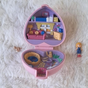 Op de afbeelding: Een vintage hartvormig poppenhuis in roze, met een open ontwerp dat een miniatuur woonruimte en een tuin laat zien. Inclusief een kleine pop en een klein dierenfiguur. Een nostalgisch speelgoed.