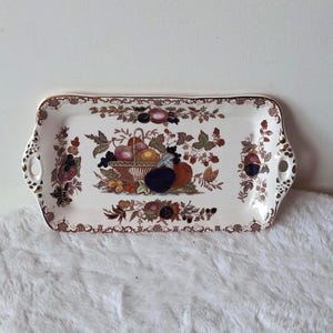 May include: A rectangular ceramic serving tray with rounded corners and integrated side handles. The white surface features a central illustration of a fruit basket surrounded by brown and gold floral borders and additional fruit motifs.