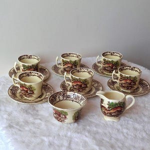 Può includere: Un set da tè in ceramica composto da cinque tazze con piattini, una zuccheriera e una lattiera. Ogni pezzo è color crema con un motivo a trasferimento marrone e verde che raffigura un paesaggio rurale con case e alberi.
