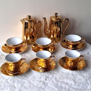 Op de afbeelding: Een theeservies bestaande uit twee metallic gouden theepotten met deksels en zes bijpassende theekopjes met schoteltjes. Elke kop-en-schotelset heeft een reflecterende gouden buitenkant en een effen witte binnenkant.