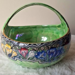 May include: A ceramic basket with a high arched handle, featuring a light green interior with a scalloped pattern. The exterior displays a band of yellow, red, white, and blue crocus flowers against a grey background, finished with gold trim.