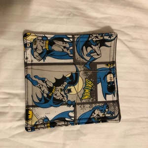 May include: A gray and blue fabric coaster with a comic book style image of Batman. The coaster has a gray background with a white distressed pattern and the word "BATMAN" in yellow letters.