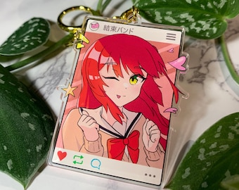 May include: A clear acrylic keychain featuring a colorful anime-style illustration of a girl with red hair and a school uniform. The keychain has a gold-colored ring and a social media-style frame with Japanese text.