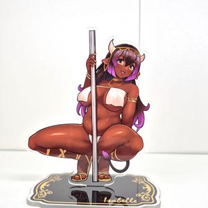 Könnte beinhalten: Ein Acryl-Aufsteller mit einer Anime-Figur mit dunkler Haut, violetten Haarspitzen und Hörnern, die an einer silbernen Stange posiert. Die Figur trägt weiße Stoffwickel und Goldschmuck. Der Sockel ist schwarz mit goldenem Filigran und dem Namen Isabelle.
