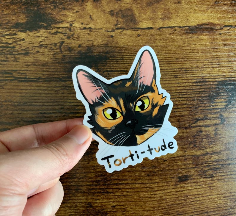 Puede incluir: Una pegatina con una ilustraci&oacute;n de dibujos animados de un gato atigrado con el texto "Torti-tude" debajo.