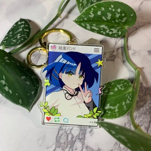 May include: A blue and white acrylic keychain featuring an anime character with blue hair and a white shirt. The character is making a peace sign with their hand. The keychain is shaped like a phone screen with a heart, a comment bubble, and three dots at the bottom. The text "結束バンド" is at the top of the screen.