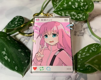 May include: A pink and blue anime character with pink hair and blue eyes is featured on a clear acrylic keychain. The character is making a peace sign with their right hand. The keychain is shaped like a phone screen with a heart, a refresh arrow, a magnifying glass, and three dots at the bottom. The text "#東京ﾊﾝﾄﾞ" is at the top of the screen.