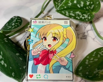May include: A clear acrylic charm featuring a cartoon girl with blonde hair and a red bow. The charm is shaped like a phone screen with a pink heart, a blue arrow, and a blue speech bubble in the bottom left corner. The text "結束バンド" is visible at the top of the charm.