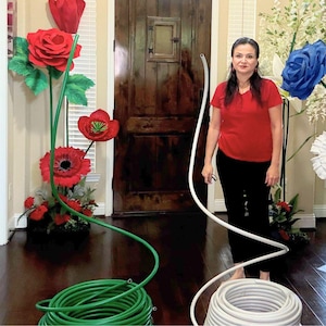 Puede incluir: Una mujer está de pie cerca de una exhibición de grandes flores de papel, incluyendo rosas rojas y amapolas, y una rosa azul. Una manguera de jardín verde está enrollada en el suelo, y otra manguera blanca es sostenida por la mujer.