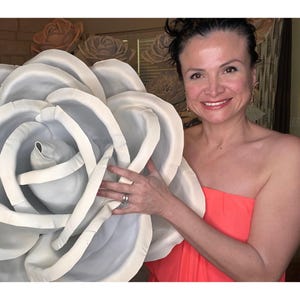 May include: A large white paper rose with a woman holding it in front of her. She is wearing a coral strapless dress.