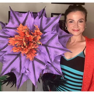 May include: A woman is holding a large purple and orange flower with black stems. The flower has many petals and is very detailed.