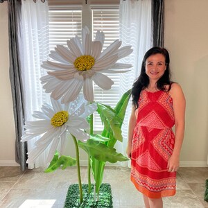 May include: A woman stands in front of a large white daisy flower arrangement. The flowers are made of paper and have green stems and leaves. The woman is wearing a red and white patterned dress.
