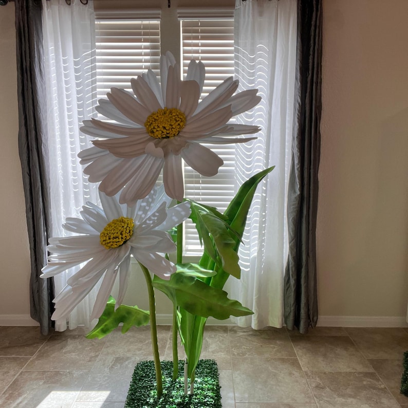 May include: Two large white daisies with yellow centers and green leaves stand in a pot with green grass. The daisies are arranged in a vase with a green base.
