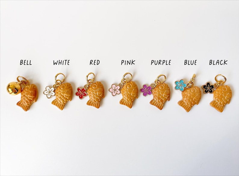 May include: A collection of Taiyaki-shaped charms in various colors, including white, red, pink, purple, blue, and black. Each charm has a gold-colored ring and a small flower or bell accent. The word "BELL" is written above the first charm.