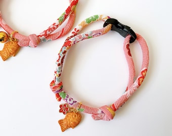 May include: Two pink and red floral patterned fabric dog collars with gold fish charms. One collar has a gold bell charm.
