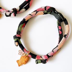 May include: Floral patterned cat collar with a black plastic buckle. The collar features a gold-colored fish charm and a small bell. The fabric has a pattern of pink, red, and green flowers on a black background.