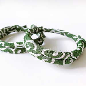 May include: Two green fabric bracelets with a white swirl pattern. The bracelets are knotted and lie on a white surface. The bracelets are a handmade accessory.